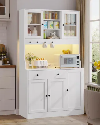 A white kitchen cabinet with glass doors and shelves, containing various items like jars, bottles, and a microwave. It is illuminated by warm LED lights.