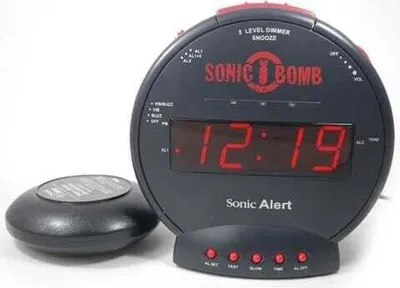 A black Sonic Alert Sonic Bomb alarm clock with a red digital display and a separate vibrating puck are shown on a white background.