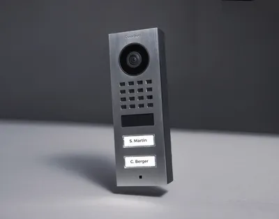 A modern, brushed stainless steel video intercom system with a camera, keypad, and two nameplates is shown against a gradient gray background.