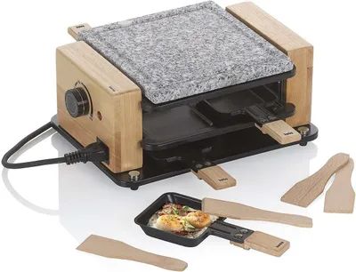 A raclette grill with a stone top and wooden accents, shown with small pans and spatulas.