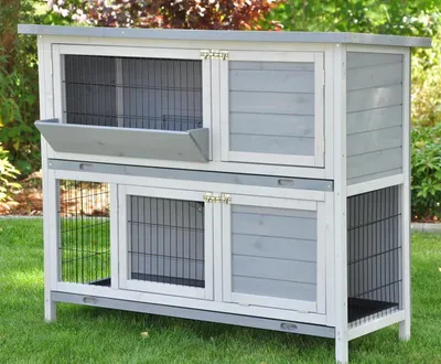 A two-tiered wooden pet cage with wire mesh doors and a ramp, set on a grassy lawn with blurred foliage in the background.