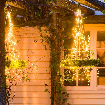 Two hanging baskets decorated with fairy lights are suspended from a wooden pergola, with a white house in the background.