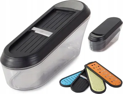 A multi-part kitchen grater set is displayed against a white background. The main grater unit, a clear plastic container with a black lid and a metal blade, is shown in the foreground. Several interchangeable grating inserts in different colors and pattern