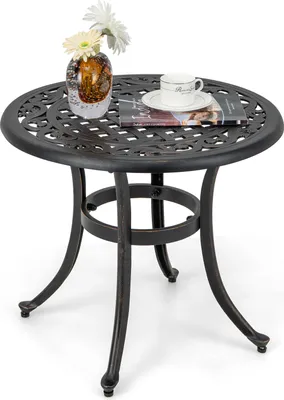 A round, dark brown cast aluminum side table with a decorative top and three curved legs, set against a white background. A glass vase with white flowers and a book with a coffee cup on top are placed on the table.