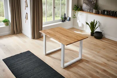 A dining table with a light wood top and white legs sits on a wooden floor with a dark rug in front of it. Large windows with beige curtains are in the background.