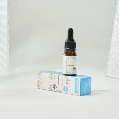 A small glass bottle with a dropper cap sits on top of its blue and white cardboard packaging, against a plain white background with subtle textured elements on the sides.