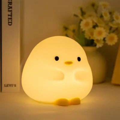 A cute, glowing chick-shaped night light sits on a surface, with a book and flowers in the background.