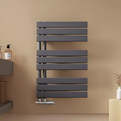 A dark gray towel radiator is mounted on a light beige wall in a bathroom setting. To the left, a gray shelf holds two soap dispensers and a woven basket. To the right, a small wooden stool holds a white vase with dried reeds.