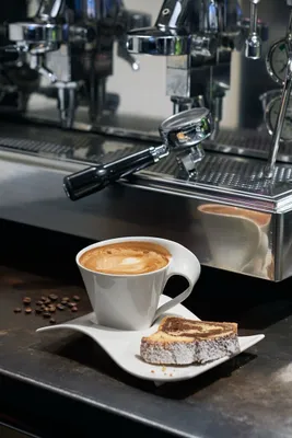 A white coffee cup with latte art and a slice of cake sits on a saucer next to a coffee machine.