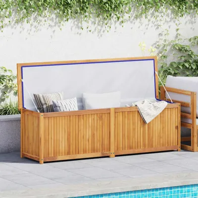 A wooden storage box with cushions and a blanket inside, placed on a patio next to a swimming pool, with greenery in the background.