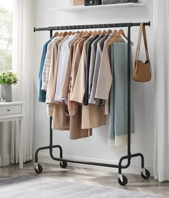 A black metal garment rack with wheels, filled with various clothing items and a blanket, placed in a room with white curtains and a dresser.
