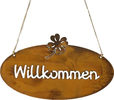 A rustic oval-shaped sign with the word "Willkommen" cut out in white letters, hanging from two strings. A small metal clover leaf decoration is attached to the top center of the sign.