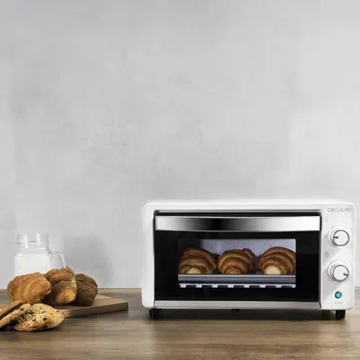 A white mini oven with croissants inside is on a wooden table, with more croissants and cookies next to it.