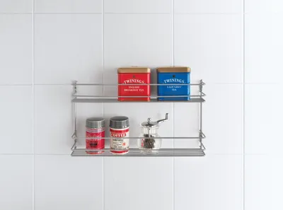 A two-tiered metal spice rack mounted on a white tiled wall, holding various spice containers and tea tins.