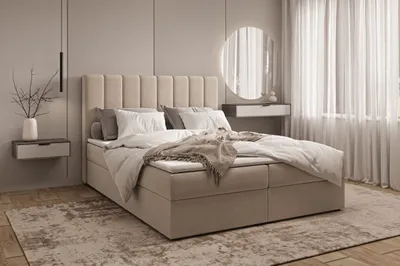 A bedroom scene featuring a large bed with a padded headboard, dressed in neutral-toned bedding and a throw blanket. A bedside table with a vase and branches is visible to the left, and sheer curtains and a round mirror are on the right.