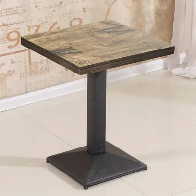 A square table with a wood-look top and a black metal base is shown in a room with a patterned wallpaper and tiled floor.