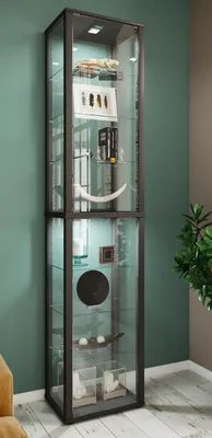 A tall, narrow glass display cabinet filled with various decorative items, standing against a teal-colored wall.