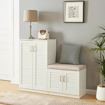 A white shoe cabinet with a built-in bench and a cushion, placed in a room with a plant and a framed picture on the wall.