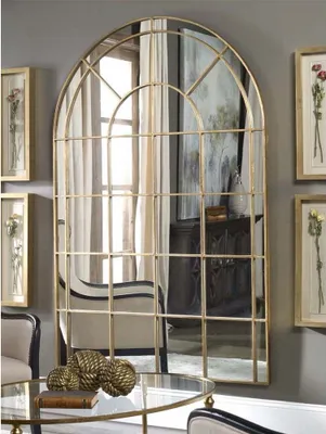 A large arched mirror with a gold frame is the focal point of a room, reflecting furniture and curtains. Decorative items are placed on a glass table in the foreground.