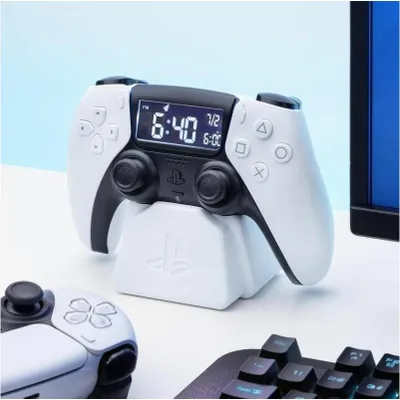 A white PlayStation PS5 controller alarm clock is displayed on a white surface, with a keyboard and a monitor in the background.