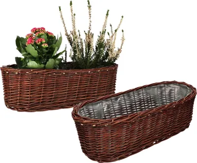 Two oval wicker planters are shown against a white background. One planter is filled with plants, including a flowering plant with pink blooms and a plant with white flowers. The second planter is empty and lined with plastic.