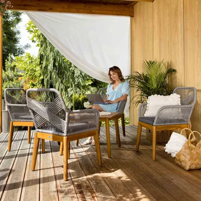 A woman is working on her laptop on a covered patio, surrounded by outdoor furniture and greenery.