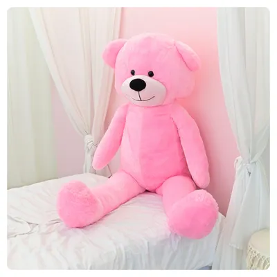 A large pink teddy bear sitting on a white bed with sheer white curtains in the background.