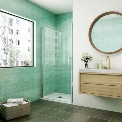 A modern bathroom with green tiled walls, a glass shower enclosure, a wooden vanity, and a round mirror.