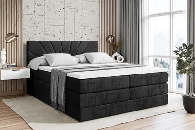 A bedroom scene featuring a large, upholstered bed with a headboard, mattress, and topper. The bed is set against a wall with textured panels and a large window with curtains. Decorative items like a plant and a vase are also visible.