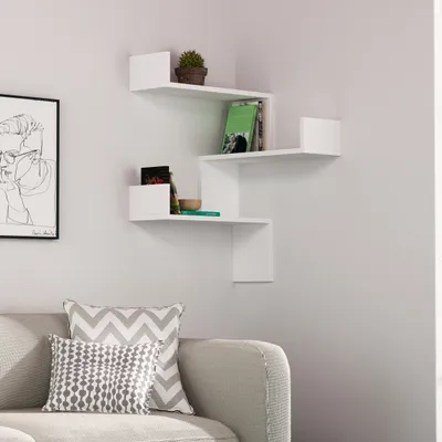 A white corner shelf unit with books and a plant displayed on it, positioned on a light gray wall above a sofa with patterned pillows.