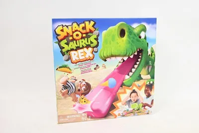 The image shows the front of the Snack-o-Saurus Rex board game box, featuring a cartoon dinosaur with its tongue extended, about to grab food items. Several children are depicted on the box, reacting to the game.