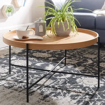 A round coffee table with a wooden top and black metal legs, set on a patterned rug in a living room. A plant, a cup of coffee, and a sugar bowl are on the table.