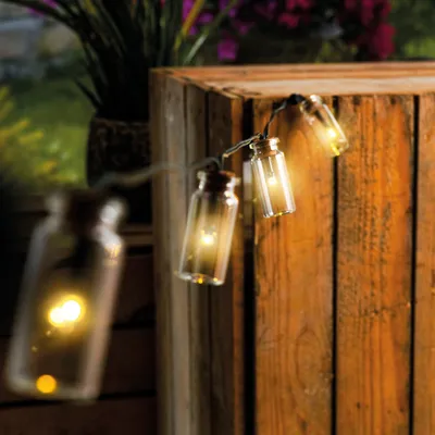 String lights with small glass bottles containing LEDs are draped over a wooden crate, with plants in the background.