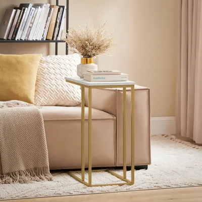 A small side table with a marble top and gold metal frame sits next to a plush sofa. Decorative items like books and a vase with dried flowers are on the table.