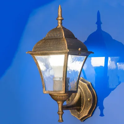 A close-up shot of a gold-colored outdoor wall lamp mounted on a blue wall, with its shadow visible.
