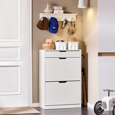 A white shoe cabinet with two drawers and a shelf above, located in an entryway next to a white door. There are hats, keys, and small baskets on the shelf.