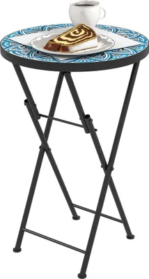 A small, round, foldable side table with a mosaic tiled top, holding a slice of cake and a cup of coffee.