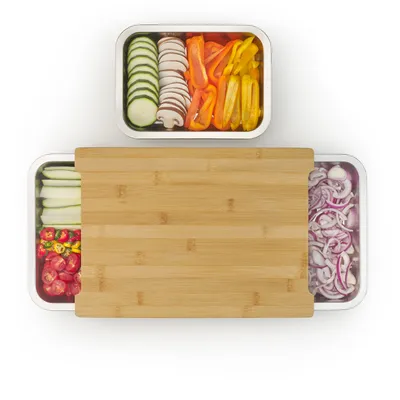 A top-down view of a bamboo cutting board with stainless steel trays filled with chopped vegetables.