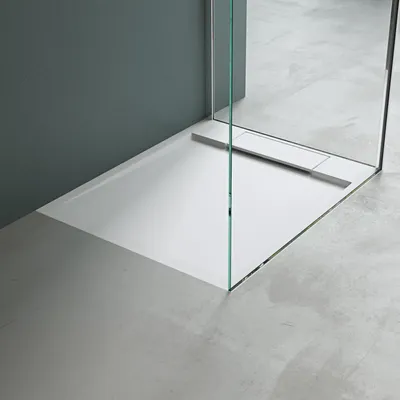 A modern shower tray with a glass partition, set against a gray wall and a concrete floor.
