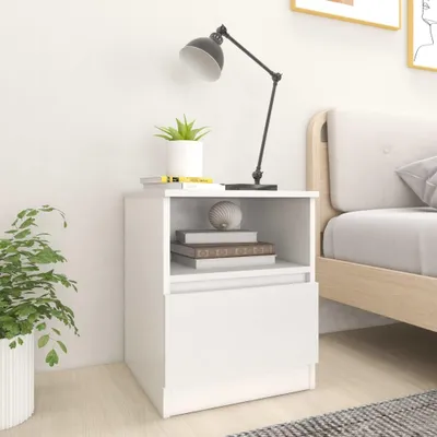 A white nightstand with a drawer and an open shelf, placed next to a bed. A lamp and a potted plant are on top of the nightstand.