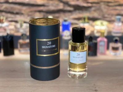 A perfume bottle and its cylindrical packaging are displayed on a wooden surface, with other perfume bottles blurred in the background.