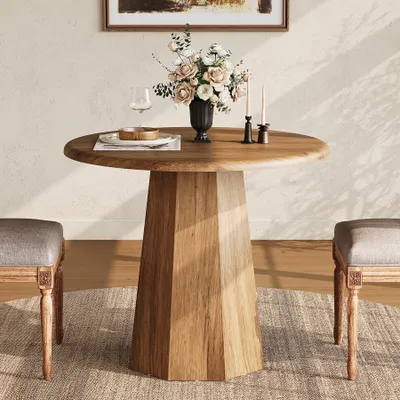 A round wooden dining table with a vase of flowers and two candles on it, placed on a rug with two chairs on either side.
