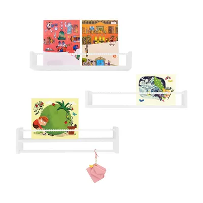 Three white wall-mounted shelves displaying children's books or artwork. A small pink origami-like charm hangs from the bottom shelf.