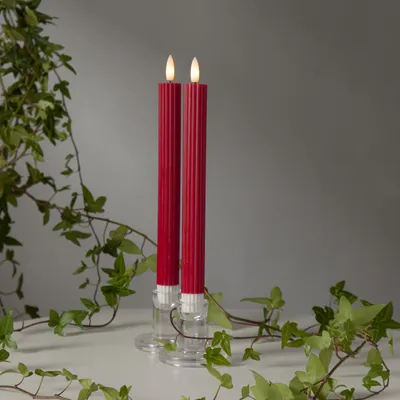 Two red LED taper candles with a flame-like top are placed in clear glass candle holders, surrounded by green ivy on a white surface against a gray background.