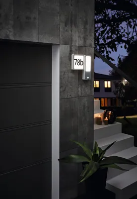 An outdoor wall lamp with house numbers is mounted on a textured wall next to a dark door. Steps with candles lead up to a house in the background.