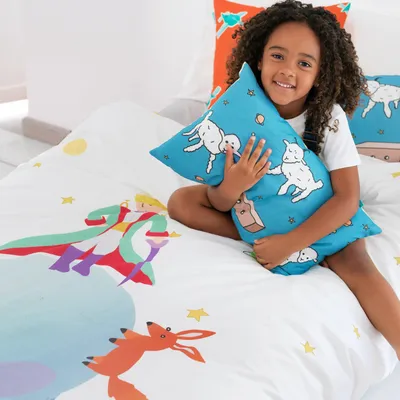 A young girl is sitting on a bed with bedding featuring illustrations from "Le Petit Prince."