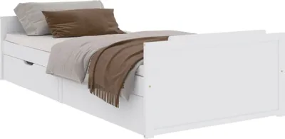 A white wooden bed with two drawers, made of pine wood, measuring 90x200 cm. The bed is shown with a light gray pillow and a brown pillow, as well as a brown blanket with fringes.