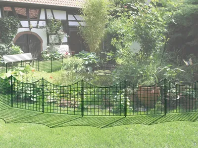 A garden scene with a fence in the foreground and a house and pond in the background.