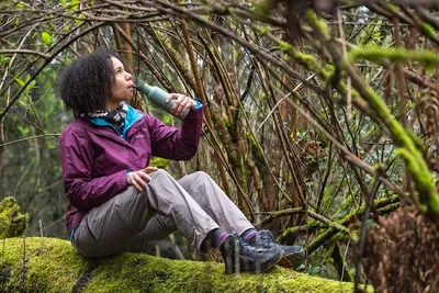 A woman sits on a moss-covered log in a forest, drinking from a water bottle.