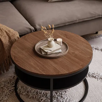 A round coffee table with a wooden top and a black storage shelf underneath, placed on a patterned rug next to a sofa.